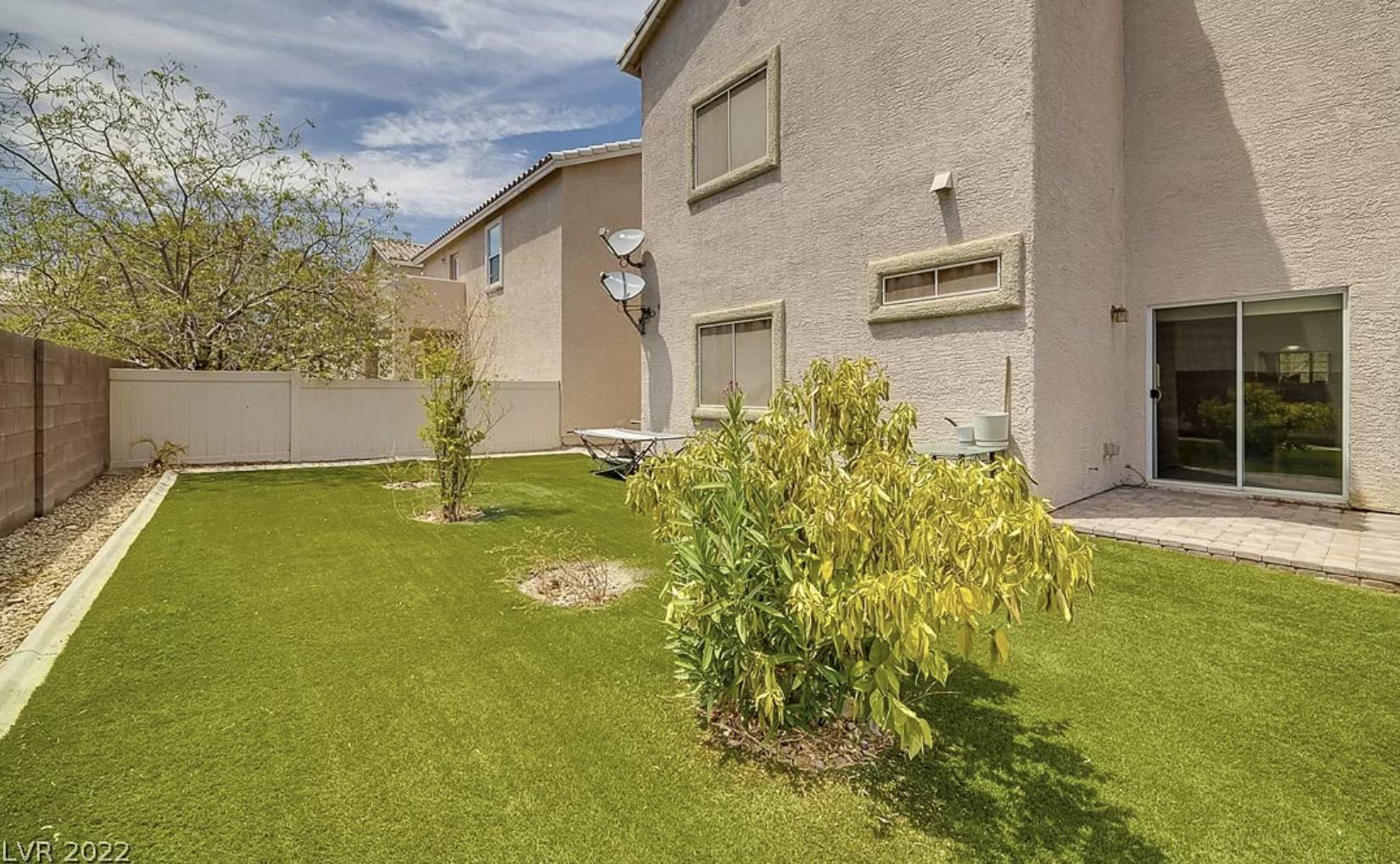 The bright green back yard of a large, beige home with several small green trees